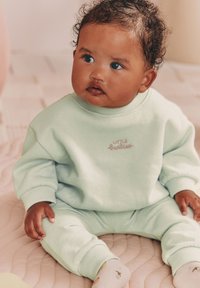 Ensemble en coton vert menthe composé d'un sweat-shirt et d'un pantalon avec poignets côtelés, détail brodé "LITTLE" et chaussettes à motifs sur une surface texturée.