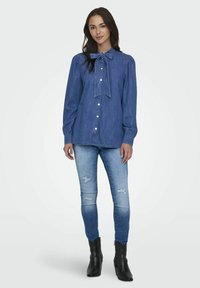 Denimskjorte med sløyfe-hals, lange ermer og knappelukking. Kombinert med slitte lyseblå jeans og svarte ankelstøvler.