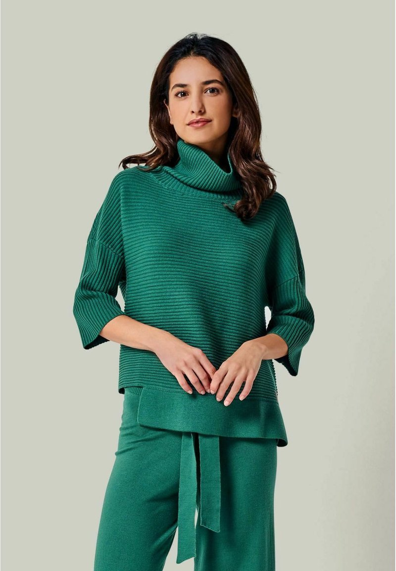 Grüner Strickpullover mit hohem, gerippter Rollkragen, 3/4-Ärmeln und entspannter Passform, kombiniert mit passenden tapered Hosen und einem Bindedetail.