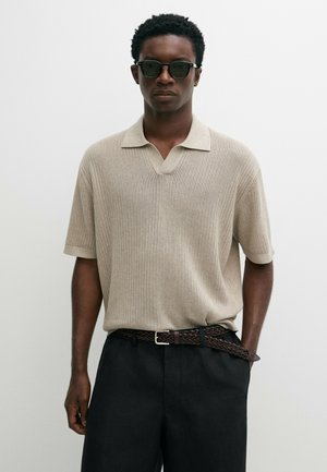 Mann trägt schwarze Sonnenbrille, beige kurzärmeliges Strick-Poloshirt, schwarze Hose und braunen geflochtenen Gürtel mit einer Hand in der Tasche.