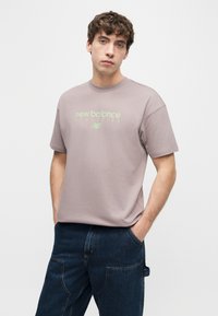 Νέος άνδρας που φοράει ανοιχτό μοβ μπλουζάκι New Balance Athletics και σκούρα μπλε τζιν, στέκεται με τα χέρια στις τσέπες μπροστά σε λευκό φόντο.