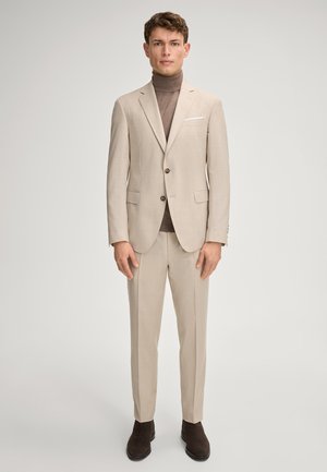 Mężczyzna w beżowym garniturze, brązowym swetrze z golfem i ciemnobrązowych butach, stojący na jednolitym jasnym tle.