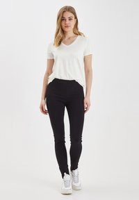 T-shirt bianco a V abbinato a pantaloni neri attillati e sneakers bianche. I pantaloni hanno una texture liscia e tasche laterali.