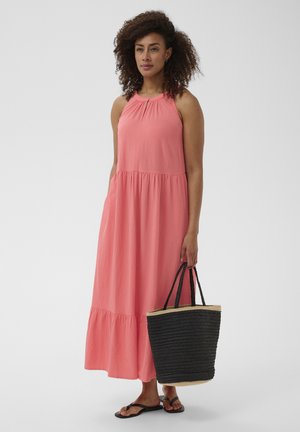 Donna con capelli ricci che indossa un maxi abito rosa senza maniche, tiene una borsa tote nera intrecciata e calza sandali neri.