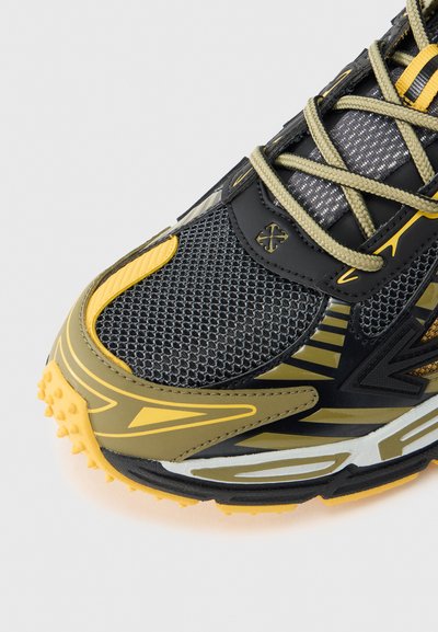 Μαύρο και κίτρινο αθλητικό παπούτσι με υφασμάτινο επάνω μέρος από υφή διχτυού, λαστιχένια σόλα με καρφιά, ανακλαστικές λεπτομέρειες και σχεδιασμένα συνθετικά επικαλύμματα.