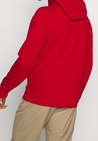Personne portant un sweat à capuche rouge et un pantalon beige, vue de dos sur un fond clair uni.