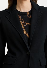 Zwarte blazer met een getailleerde pasvorm, geknipte revers en een enkele knoopsluiting. Daaronder een gestreept shirt met gouden kettingpatronen.