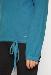 Un pull turquoise avec un cordon de serrage à l'ourlet, confectionné dans un tissu doux. Les manches longues présentent des bords-côtes et un détail de logo.