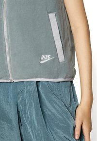 Grå fleeceväst med dragkedja fram, har två sidofickor, grå kant och en vit Nike-logotyp längst ner till höger.
