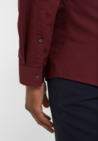 Bordeaux shirt met knoopmanchetten en gestikte zoom, gecombineerd met donkerblauwe broek. Close-up van de details van de pols en mouw zichtbaar.