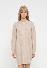 Ξανθιά γυναίκα που φοράει ένα μπεζ φόρεμα με πλέξη καλωδίου και μακρύ μανίκι, στέκεται μπροστά σε λευκό φόντο.