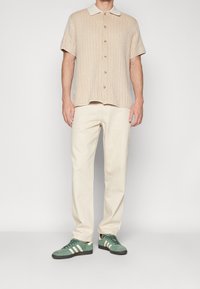 Camisa de botones de manga corta en beige claro con textura acanalada, pantalones cremosos y zapatillas de ante verde con rayas blancas.