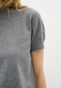 Nahaufnahme einer Person, die einen kurzärmligen grauen Strickpullover vor einem einfarbigen hellen Hintergrund trägt, wobei Schulter und Oberarm sichtbar sind.