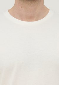 Κοντινό πλάνο του λαιμού και του άνω μέρους του στήθους ενός ατόμου που φοράει απλό λευκό T-shirt με στρογγυλή λαιμόκοψη, μπροστά σε ένα φωτεινό φόντο.