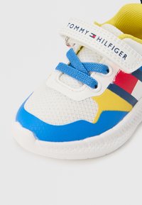 Biely atletický topánok s modrými, žltými a červenými akcentmi. Sieťovaný zvršok, gumová podrážka, modré šnúrky a nastaviteľný pásik s logom &quot;TOMMY HILFIGER&quot;.