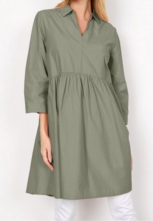 Femme portant une robe tunique ample à manches longues vert olive, avec col et taille froncée, associée à un pantalon blanc sur un fond uni.