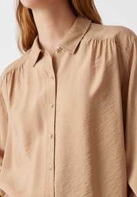 Camicia beige con colletto, in tessuto texturizzato e con dettagli arricciati sulle spalle, dotata di piccoli bottoni lungo la parte anteriore.