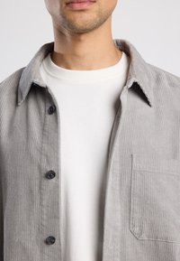 Gray corduroy shirt with a button-down front and chest pocket, layered over a white crew-neck t-shirt. Soft texture with vertical ribbing.