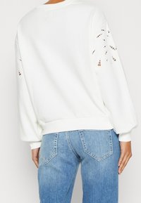 Bílá mikina s uvolněným střihem, zdobená výseky na rukávech. V kombinaci s světle modrými džíny s tapered nohavicemi.