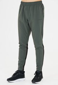 Mäns träningsbyxor i mörkgrön, tillverkade av lättviktig tyg med elastisk midja och svarta accentpartier på sidorna. Kombineras med svarta sneakers.