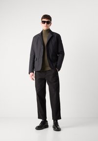 Chaqueta texturada negra sobre un suéter de cuello alto verde oliva, combinada con pantalones negros y zapatos negros con cordones, finalizada con gafas de sol oscuras.