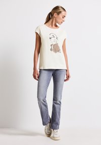 Camiseta corta de manga de color crema con una ilustración impresa en el frente. Combinada con jeans rectos de color azul claro y zapatillas neutras.