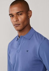 Pánska polokošeľa s dlhým rukávom z ľahkej modrej bavlny, s dvoma gombíkmi na zapínanie a kontrastným čiernym logom na hrudi. Hladká textúra.