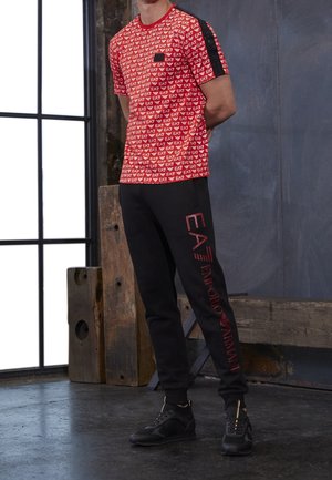 Homme portant un t-shirt rouge avec logo EA7 imprimé et un pantalon de survêtement noir avec texte rouge EA7 Emporio Armani, debout à l'intérieur.