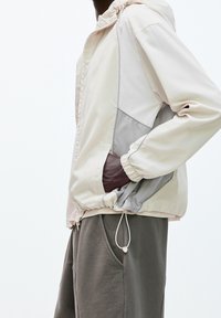 Person som bär en ljus beige och grå vindjacka med handen i fickan, tillsammans med lösa mörkgrå byxor, mot en neutral vit bakgrund.