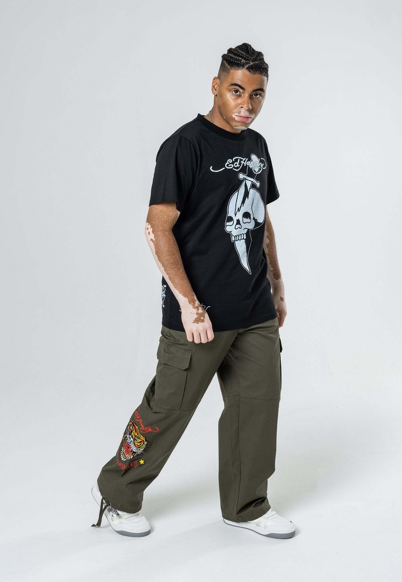Czarna koszulka z nadrukiem czaszki i tekstem; oliwkowe spodnie cargo z wyszytym tygrysem; białe sneakersy; prosta postawa.