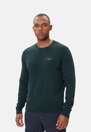 Uomo che indossa un maglione verde scuro a maniche lunghe con un piccolo emblema ricamato sul petto, in piedi davanti a uno sfondo semplice.