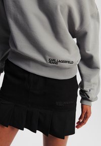 Sudadera gris con dobladillo acanalado, que presenta "KARL LAGERFELD JEANS" en bordado negro. Falda corta plisada negra con un detalle de logo similar.