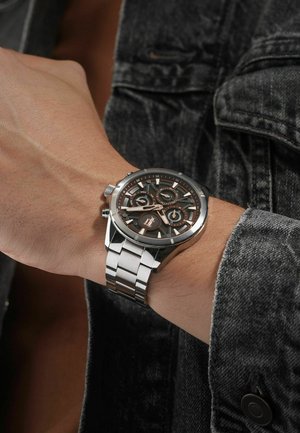 Silver stainless steel watch with a black dial, three sub-dials, and glossy accents. Worn on a wrist, featuring a linked bracelet design.