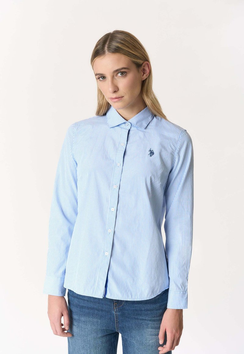 Donna bionda con capelli lisci che indossa una camicia a righe azzurre e bianche con bottoni e jeans blu, in piedi davanti a uno sfondo bianco.