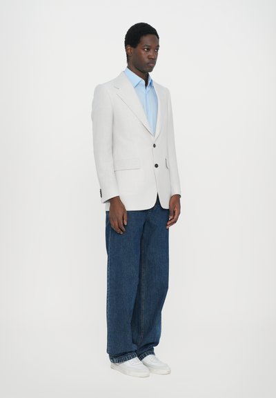Blazer gri deschis, cămașă albastră, blugi șaici închis și adidași albi purtați de o figură în picioare. Butoane notabile pe blazer.