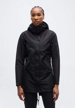 Γυναίκα με πλεγμένα μαλλιά που φοράει μαύρο αδιάβροχο με κουκούλα και εμπρόσθιες τσέπες με κουμπιά, στέκεται μπροστά σε ένα απλό φωτεινό φόντο.