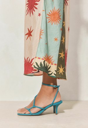 Bare foot wearing turquoise strappy kitten heel sandal, ankle strap buckled, next to colorful sun and star patterned flowy skirt.