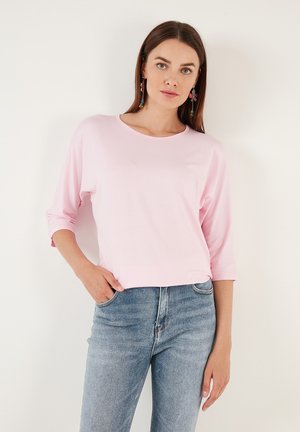 Lyserød, langærmet top med rund halsudskæring og opsmøgte ærmer, parret med lyseblå jeans. Glat stofstruktur.