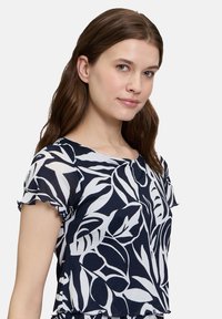Granatowa bluzka z białym wzorem liści, krótkie rękawy, okrągły dekolt i falbaniasty dół. Lekki materiał o gładkiej teksturze.