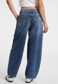 Weite blaue Jeans aus Denim mit verblasster Optik, ausgestattet mit zwei Gesäßtaschen und einem schmalen Knöchelbereich. Getragen mit weißen Sportschuhen.