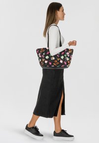 Donna che cammina indossando una gonna nera, una maglietta a maniche lunghe bianca, scarpe da ginnastica nere, e porta una grande borsa a tracolla nera intrecciata con motivo floreale.