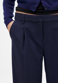 Navy-Slacks mit Falten, seitlichen Taschen und einer glatten Stofftextur. Kombiniert mit einem dunklen, figurbetonten Oberteil und einem lila Cardigan.