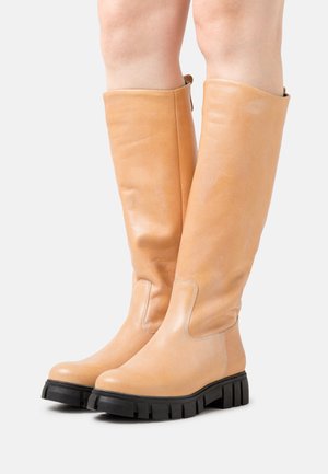 Botas de joelho de couro castanho com uma textura suave, bico arredondado e uma sola preta de borracha robusta. Detalhes da costura lateral são visíveis.