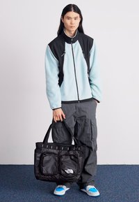 Casaco de fleece azul claro com detalhes pretos, calças cargo cinza e uma mala tote preta da North Face, com dois bolsos frontais e al�ças.