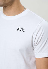 Mand iført hvid T-shirt med lille sort logo af to siddende figurer på øverste venstre bryst, synlig sølvkæde.