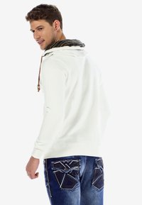 White hoodie with a patterned black and gold lined hood, featuring a drawstring and a slim fit. Paired with blue jeans with decorative back pockets.