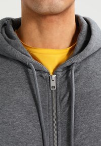 Sudadera gris con cremallera frontal, capucha con cordones y tejido suave y texturizado, sobre una camiseta amarilla debajo.