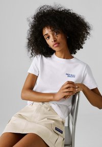 Γυναίκα με σγουρά μαλλιά, φορώντας λευκό T-shirt της Red Bull Racing και μπεζ φούστα, κάθεται σε μεταλλική καρέκλα μπροστά σε γκρι φόντο.