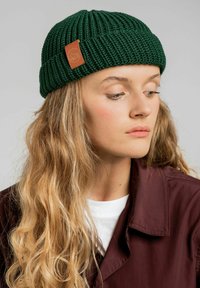 Zielona dziana czapka z prążkowaną teksturą i brązow ą skórzaną metką z logo, noszona powyżej bordowej kurtki i białego topu.