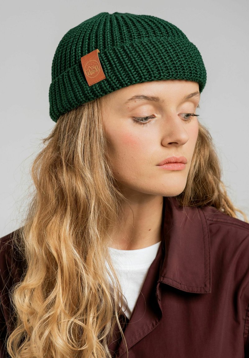 Zielona dziana czapka z prążkowaną teksturą i brązową skórzaną metką z logo, noszona powyżej bordowej kurtki i białego topu.